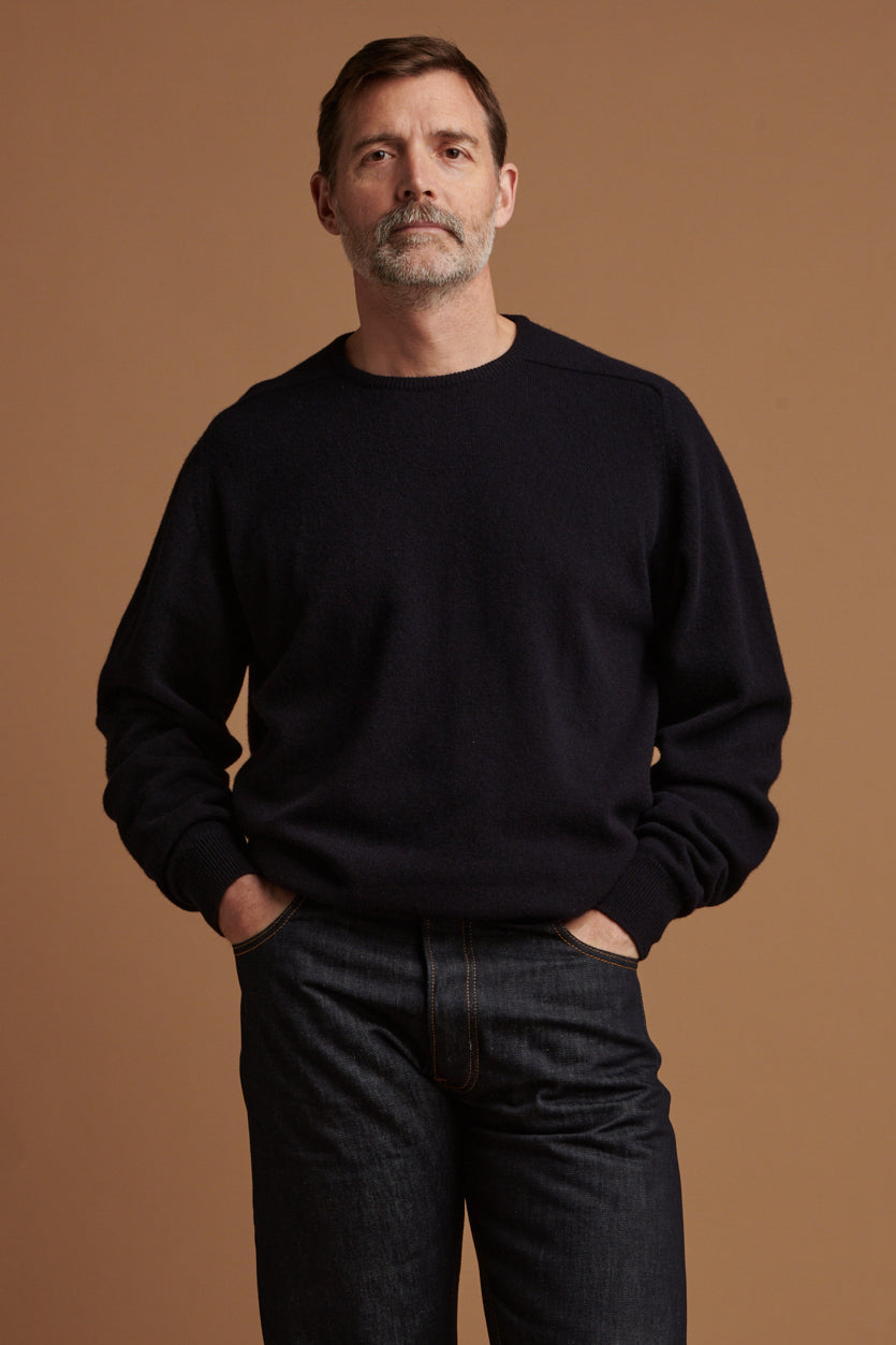 Thigh up front of Patrick Grant wearing lambswool saddle shoulder crew neck jumper in navy, worn with loose fit button fly Arran jeans in indigo
